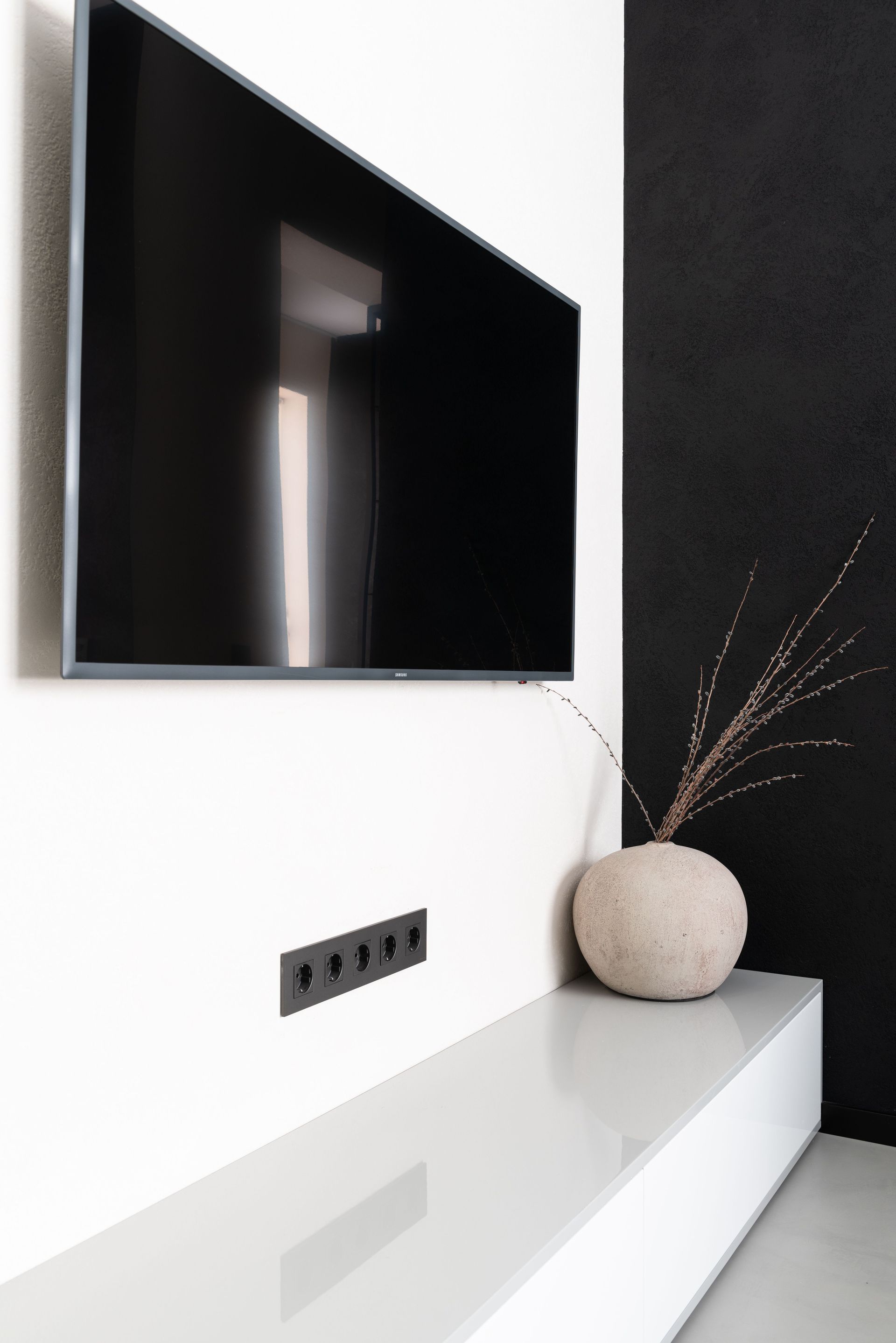 Modern television set hanging on white wall above round vase with branches in stylish apartment with black screen